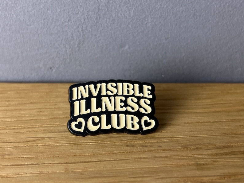 May include: A black and white enamel pin with the words "INVISIBLE ILLNESS CLUB" in a bold, stylized font. The pin features two heart shapes and a black border. The pin is on a wooden surface.