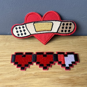 Könnte beinhalten: Zwei gestickte Bügelbilder. Das erste Patch ist ein rotes Herz mit einem weißen Verband. Das zweite Patch ist eine pixelartige Herz-Gesundheitsleiste mit drei roten Herzen und einem grauen Herzen.