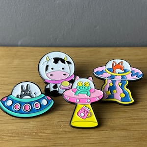 Könnte beinhalten: Satz mit fünf Emaille-Pins mit Cartoon-Tieren in Raumschiffen. Die Pins sind bunt und zeigen eine Kuh, einen Fuchs, einen Frosch und eine Totoro-ähnliche Kreatur. Die Pins sind perfekt, um Ihrem Rucksack, Ihrer Jacke oder Ihrer Tasche einen Hauch von Verspieltheit zu verleihen.