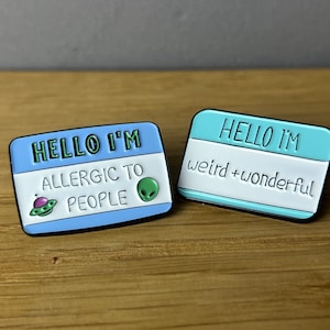 Könnte beinhalten: Zwei blaue und türkisfarbene Emaille-Pins in Form von Namensschildern. Der obere Pin lautet "Hello I'm allergic to people" mit einer kleinen Alien- und Raumschiffgrafik. Der untere Pin lautet "Hello I'm weird + wonderful".