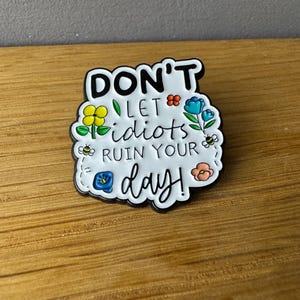 Könnte beinhalten: Ein weiß-schwarzer Emaille-Pin mit dem Schriftzug "DON'T LET idiots RUIN YOUR day!" umgeben von bunten Blumen und Bienen. Der Pin befindet sich auf einer Holzoberfläche.