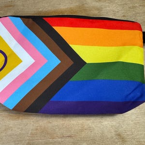 Könnte beinhalten: Eine rechteckige Tasche mit einem schwarzen Reißverschluss und einer kleinen schwarzen Schlaufe an der Seite. Die Tasche hat ein farbenfrohes Design mit einem Regenbogen und einer progressiven Pride-Flagge.