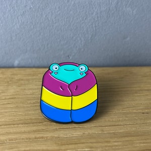 Könnte beinhalten: Ein farbenfroher Emaille-Pin mit einem stilisierten Frosch-Design. Der Frosch ist in eine Decke in den Farben der Pansexuell-Pride-Flagge gehüllt: Pink, Gelb und Blau. Der Frosch hat große, runde Augen und einen lächelnden Ausdruck.