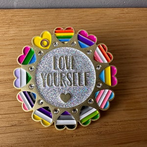 Könnte beinhalten: Goldene Anstecknadel mit Glitzerhintergrund und dem Text "LOVE YOURSELF", umgeben von einem Kreis aus bunten Herzen. Jedes Herz zeigt eine andere Farbkombination, die die LGBTQ+ Pride Flagge repräsentiert.