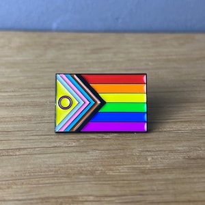 Könnte beinhalten: Rechteckiger Emaille-Pin mit dem Design der Progress Pride Flagge. Der Pin hat einen schwarzen Rand und enthält die traditionellen Regenbogenstreifen sowie die Farben der Transgender-Flagge und das Intersex-Symbol.