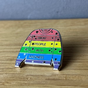 Könnte beinhalten: Eine regenbogenfarbene Emaille-Anstecknadel in Form eines Pullovers mit den Worten "Treat People With Kindness" und schwarzen Sternen.