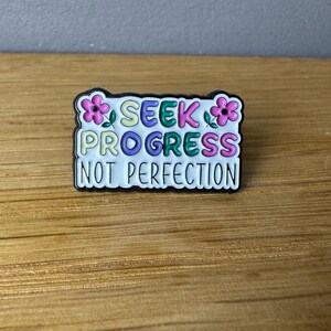 Könnte beinhalten: Rechteckiger Anstecker mit dem Text "SEEK PROGRESS NOT PERFECTION" in bunten Buchstaben. Der Anstecker hat einen schwarzen Rand und ist mit rosa Blumen verziert. Der Anstecker liegt auf einer Holzoberfläche.