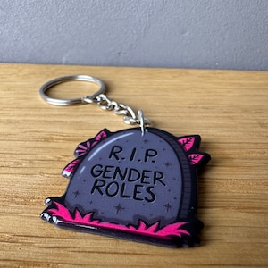 Könnte beinhalten: Ein grauer Grabstein-Schlüsselanhänger mit rosa Blumen und dem Text "R.I.P. Gender Roles".