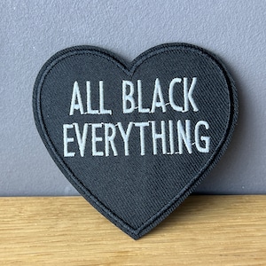 Könnte beinhalten: Schwarzer herzförmiger Patch mit weißer Stickerei, auf dem "ALL BLACK EVERYTHING" steht.