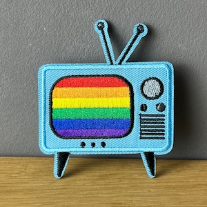 Könnte beinhalten: Gestickter Aufnäher mit einem hellblauen Retro-Fernseher mit Regenbogenfarbenem Bildschirm. Der Aufnäher hat schwarze Antennen, Beine und Details. Ein farbenfrohes und lebendiges Accessoire für Kleidung oder Taschen.