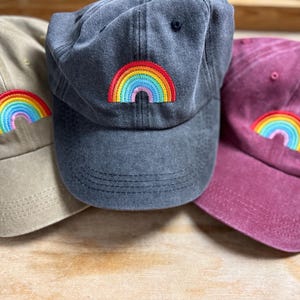 Könnte beinhalten: Drei Baseballkappen in Beige, Anthrazitgrau und Burgunderrot. Jede Kappe hat einen bunten, gestickten Regenbogen auf der Vorderseite. Die Kappen sind aus einem weichen, strukturierten Stoff gefertigt und haben einen gebogenen Schirm.