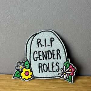 Könnte beinhalten: Ein grauer gestickter Aufnäher mit einem Grabstein-Design. Der Grabstein trägt die Aufschrift "RIP Gender Roles" mit Blumen auf beiden Seiten.