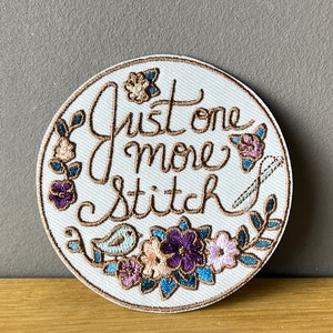 Könnte beinhalten: Ein runder weißer gestickter Aufnäher mit braunen Stichen, auf dem "Just one more stitch" steht, umgeben von einem Blumendesign mit blauen, rosa und violetten Blumen und einem kleinen blauen Vogel.