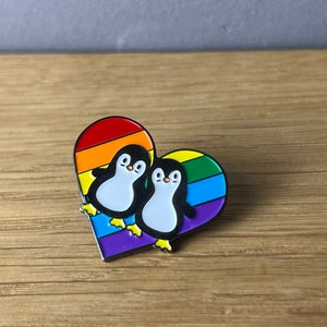 Könnte beinhalten: Ein herzförmiger Emaille-Pin mit zwei Cartoon-Pinguinen, die sich umarmen. Das Herz ist in Regenbogenstreifen unterteilt. Die Pinguine sind schwarz und weiß mit gelben Füßen. Der Pin ist auf einer Holzoberfläche platziert.