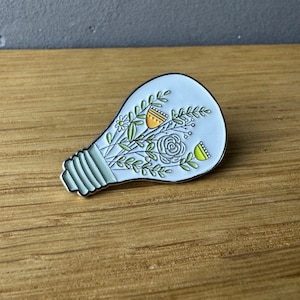 Könnte beinhalten: Eine weiße Glühbirnen-Emaille-Anstecknadel mit einem grünen und gelben Blumendesign im Inneren. Die Anstecknadel befindet sich auf einer Holzoberfläche.