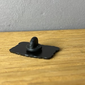 May include: A black enamel pin with a rounded rectangular shape. The pin has a black clutch back. The pin is on a wooden surface.