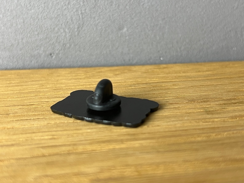 May include: A black enamel pin with a rounded rectangular shape. The pin has a black clutch back. The pin is on a wooden surface.
