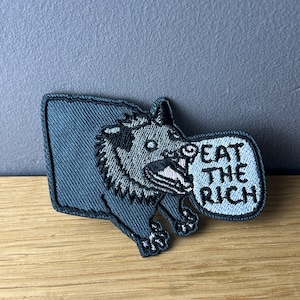 Könnte beinhalten: Ein gestickter Aufnäher in Schwarzweiß, der ein Cartoon-Opossum mit einer Sprechblase zeigt, auf der "Eat the Rich" steht.