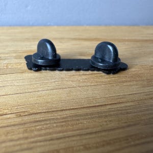 May include: A black enamel pin back with two rounded posts. The pin back is rectangular with a decorative edge. The pin is designed to be attached to fabric or other materials.
