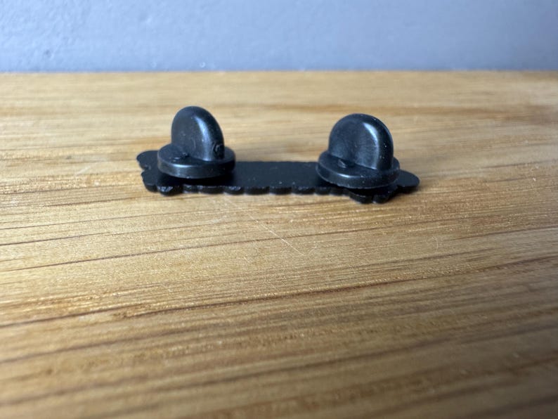 May include: A black enamel pin back with two rounded posts. The pin back is rectangular with a decorative edge. The pin is designed to be attached to fabric or other materials.