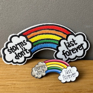 Könnte beinhalten: Set aus Regenbogen-Patches und einem Ansteck-Pin. Das Regenbogen-Design umfasst die Farben Rot, Orange, Gelb, Grün, Blau und Lila. Die Texte "storms don't" und "last forever" sind enthalten. Der Pin ist eine kleinere Version des Patches.