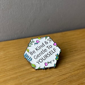Könnte beinhalten: Weißer Emaille-Pin in sechseckiger Form mit schwarzem Rand. Der Pin zeigt ein Blumenmuster mit bunten Blumen und Blättern, die den Text "Be Kind & Gentle To YOURSELF" umgeben.