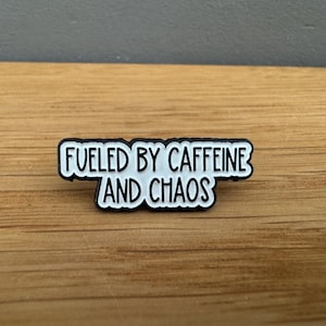 Könnte beinhalten: Weißer Emaille-Pin mit schwarzem Umriss, mit dem Text "Fueled by Caffeine and Chaos".