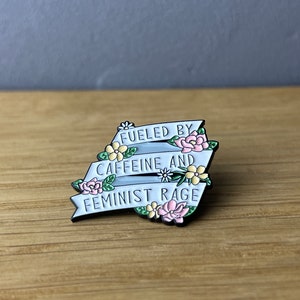 Könnte beinhalten: Metall-Anstecknadel mit Banner-Design. Das hellblaue Banner trägt die weißen Worte "FUELED BY CAFFEINE AND FEMINIST RAGE". Die Anstecknadel ist mit rosa und gelben Blumen und grünen Blättern verziert.