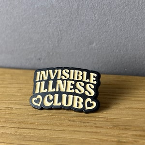 May include: A black and tan pin with the words "INVISIBLE ILLNESS CLUB" in a bold, stylized font. The pin features two heart shapes. The pin is on a wooden surface.