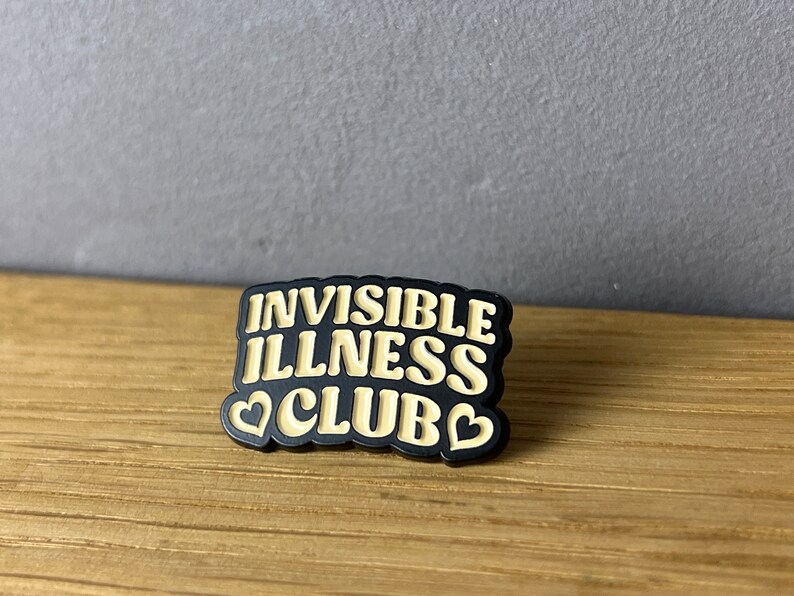 May include: A black and tan pin with the words "INVISIBLE ILLNESS CLUB" in a bold, stylized font. The pin features two heart shapes. The pin is on a wooden surface.