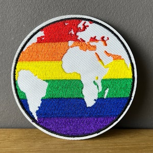 Könnte beinhalten: Ein runder gestickter Aufnäher mit einem weißen Umriss und einem weißen Weltkarten-Design. Die Weltkarte ist von einem Regenbogen aus Farben umgeben, darunter Rot, Orange, Gelb, Grün, Blau, Indigo und Violett.