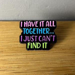 Könnte beinhalten: Ein schwarzer rechteckiger Anstecker mit dem Text "I HAVE IT ALL TOGETHER... I JUST CAN'T FIND IT" in Pink, Blau, Lila und Grün. Der Anstecker liegt auf einer Holzoberfläche.