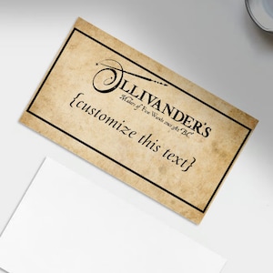 Pode incluir: Um cartão de visita com um design de pergaminho vintage, com o texto "Ollivander's Makers of Fine Wands since 382 BC" e "{customize this text}". Uma caneta rosa dourada, uma régua e um pequeno cartão branco também são visíveis.
