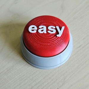 May include: A red button with the word "easy" in white letters. The button has a ridged surface and sits in a gray base. The button is on a light-colored wooden surface.