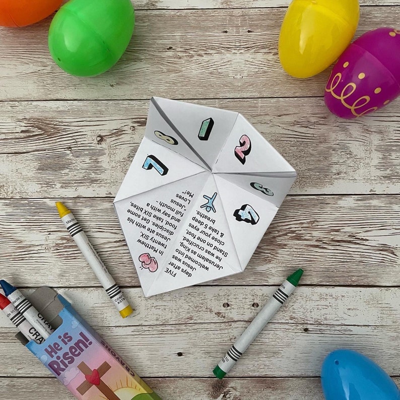 May include: A white paper fortune teller with colourful numbers and illustrations. The fortune teller is open and shows phrases like "Jesus loves me!" and "Stand on one foot, close your eyes and take 5 deep breaths". The fortune teller is on a white wooden surface with colourful Easter eggs.