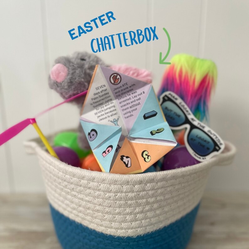 May include: A white and blue woven basket filled with Easter themed items, including a colourful origami fortune teller, a pink and yellow toy, a green and yellow toy, a purple toy, and a pair of sunglasses with a "Keep the Outtihne" sticker. The basket is labelled "Easter Chatterbox".