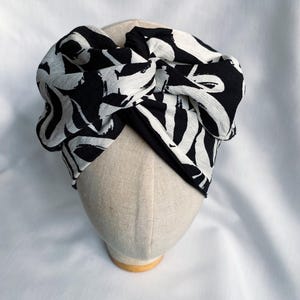 May include: A wide fabric headband featuring a bold black and white abstract brushstroke pattern. The headband is designed with a central twisted knot detail at the top, creating a gathered, voluminous appearance across the front of the head.
