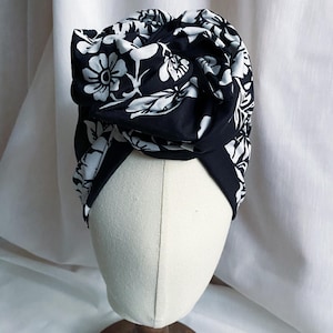May include: A black and white floral print head wrap featuring a large, knotted fabric detail at the front. The fabric is patterned with white flowers and leaves against a solid black background, wrapped securely around a neutral-coloured mannequin head.