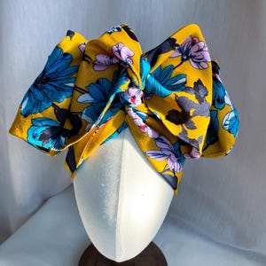 May include: A mustard yellow fabric headband featuring a large, knotted bow detail. The headband is decorated with a vibrant floral pattern consisting of bright blue, light purple, and dark grey flowers with green stems and leaves across the surface.