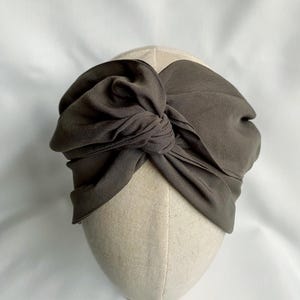 May include: A dark olive green knotted headband. The fabric is draped and tied into a large bow on top of the head. The headband is displayed on a mannequin head with a neutral fabric.