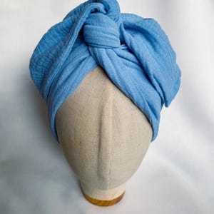 May include: A light blue turban-style headwrap made of a textured, crinkled fabric. The headwrap features a prominent central knot detail at the front, with soft, gathered folds that wrap around the head, creating a voluminous and layered appearance.