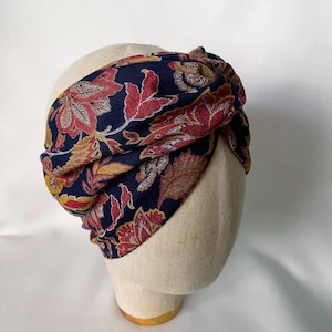 May include: A wide fabric headband featuring a dark navy blue base with a repeating floral pattern in shades of red, gold, and cream. The headband has a twisted knot detail at the front and is displayed on a neutral-coloured mannequin head.