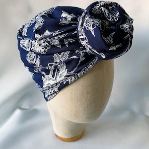 May include: A navy blue turban with a white floral pattern is displayed on a mannequin head. The turban is made of soft fabric and features a large, decorative knot on top. The floral design includes detailed outlines of leaves and flowers.