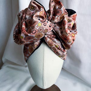 May include: A fabric headwrap featuring a repeating pattern of circular motifs in shades of pink, brown, and gold. The headwrap is tied in a large, voluminous bow at the top, with a black fabric lining visible along the edges of the folded material.