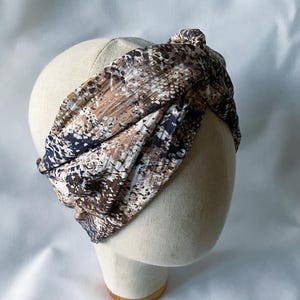 May include: A patterned headband in shades of brown, beige, and navy blue, displayed on a mannequin head. The fabric has a textured appearance and is knotted at the top. The headband is designed to be worn around the head.