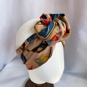 May include: A wide fabric headband featuring a tan base colour with a printed pattern of abstract leaves in shades of dark blue, mustard yellow, and bright orange. The headband is styled with a large, knotted fabric bow detail at the top centre.