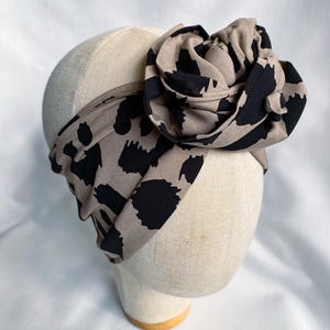 May include: A wide fabric headband featuring a beige base with an irregular black animal print pattern. The headband includes a large, gathered fabric flower accent on the side, constructed from the same patterned material as the band itself.