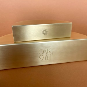 Pu&ograve; includere: Due scatole di metallo dorato impilate l'una sull'altra. La scatola superiore ha il testo "OAK and 9TH" inciso sul davanti. La scatola inferiore ha lo stesso testo inciso sul davanti.