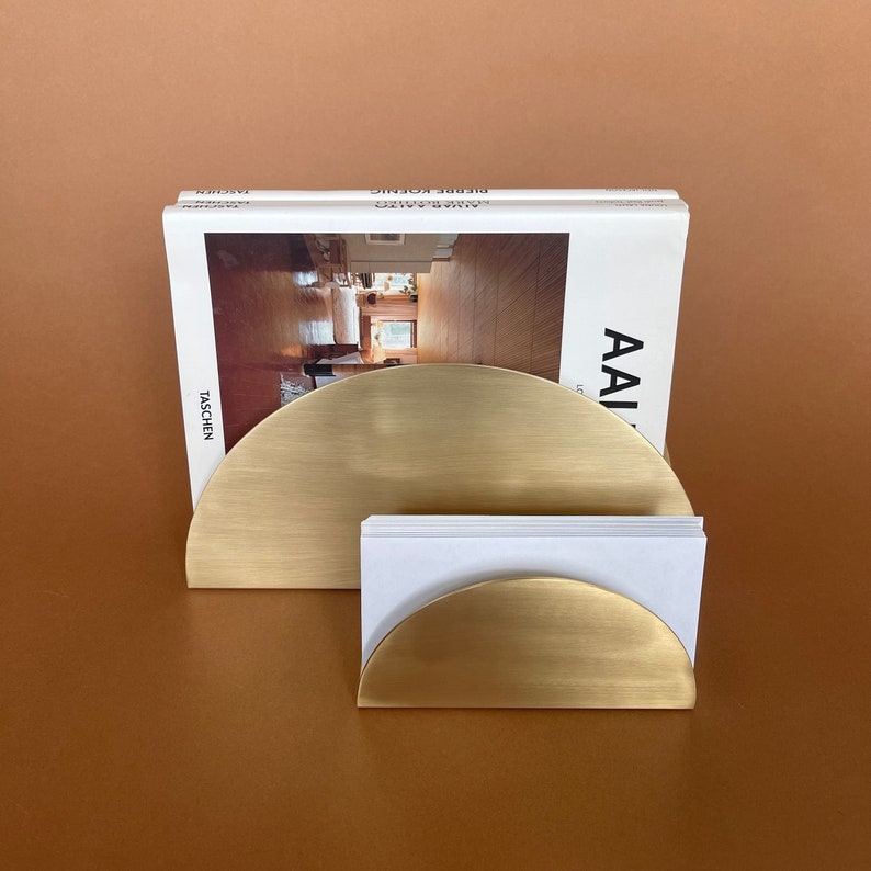 Pu&ograve; includere: Un organizzatore da scrivania in metallo dorato con due scomparti semicircolari. Lo scomparto pi&ugrave; grande contiene un libro intitolato "Alvar Aalto" di Gilbert Rohde, e lo scomparto pi&ugrave; piccolo contiene un mazzo di biglietti da visita bianchi.