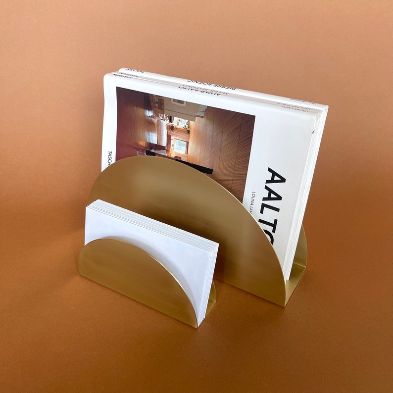 Pu&ograve; includere: Un porta riviste e lettere in metallo dorato con due scomparti semicircolari. Il porta riviste contiene una pila di libri bianchi con il testo "AALTO" sulla copertina.
